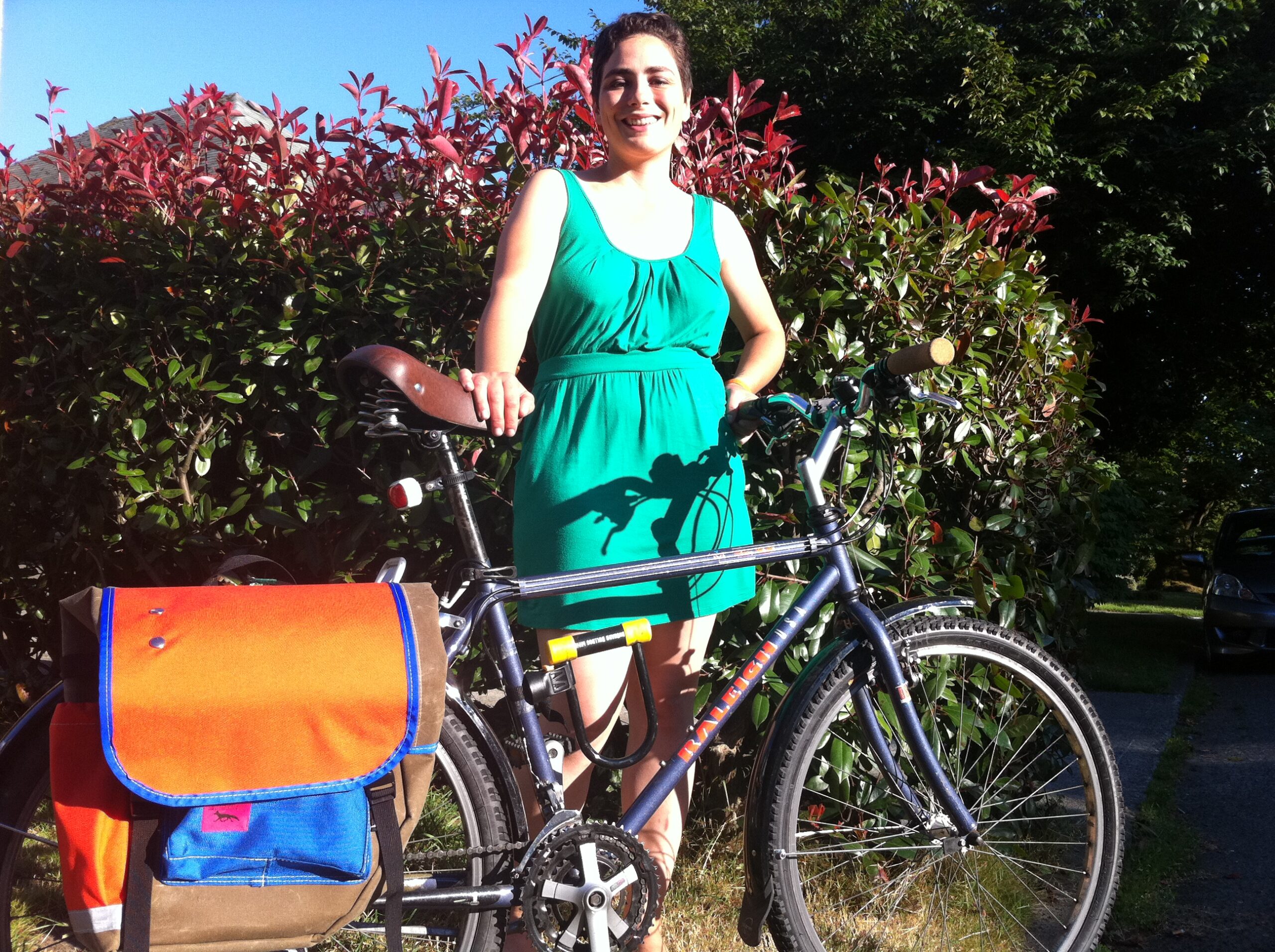 Catherine in a green dress posing with her purple bicycle and custom Swift pannier in front of a bush on a sunny day.