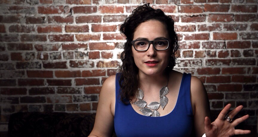 Catherine wearing glasses and a blue dress and about to speak in front of a brick wall.